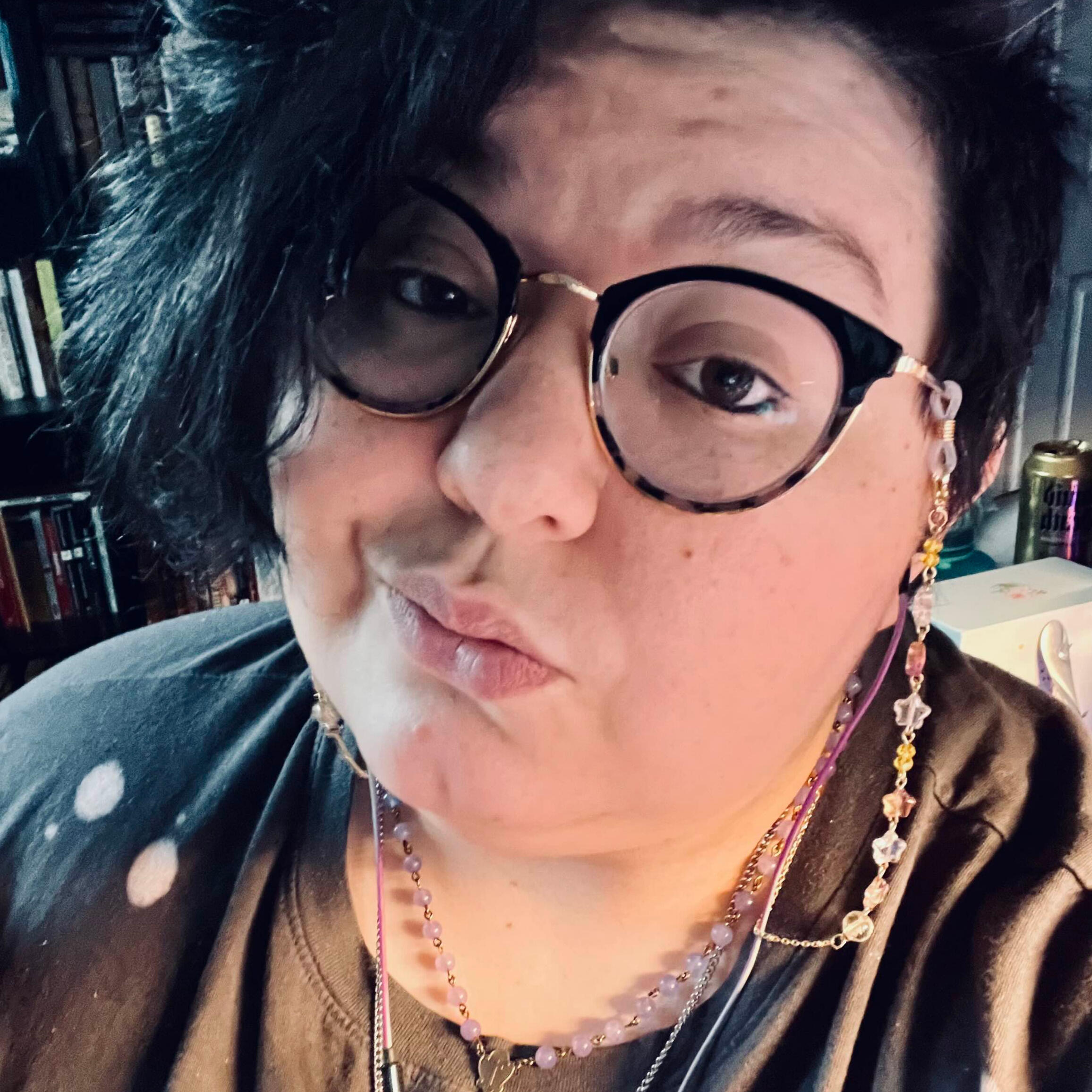 a headshot of quinn smoldering into the camera. arms crossed over their chest, their chin rests on the back of one hand. their hair is up in a high bun and they wear a wine red tank top underneath black overalls. they wear thick, clear-framed glasses, and